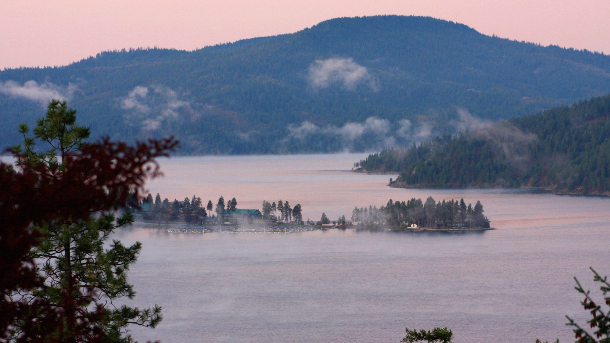 Morning mist over Coeur d'Alene Lake at dawn, with forested hills and a wooded point